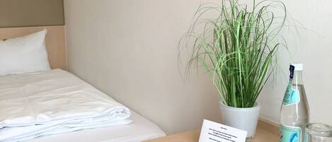Una habitación de hotel con una cama, una planta en maceta, una botella de agua y un vaso sobre una mesa de madera.