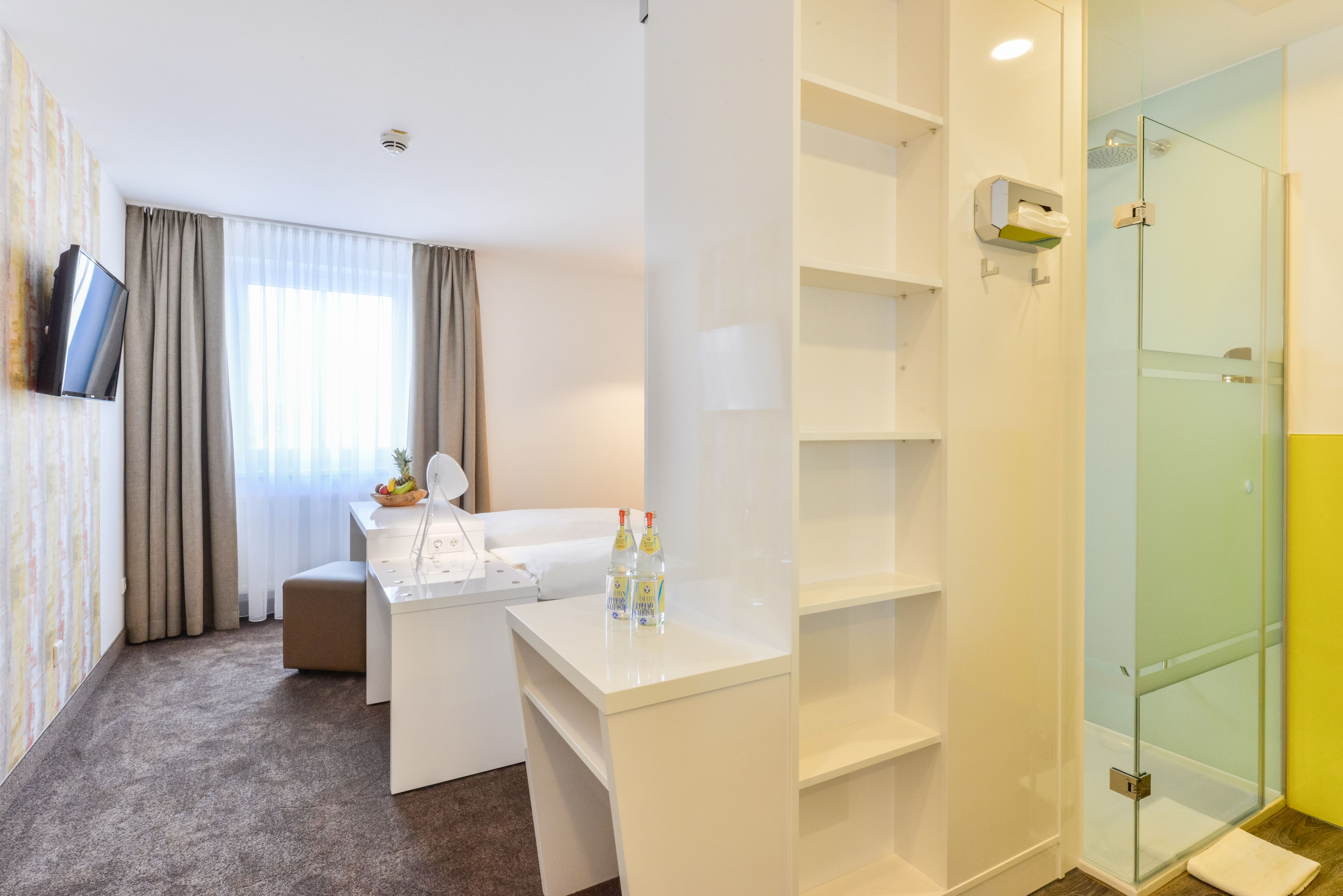 A hotel room with a white desk, a TV mounted on the wall, a glass shower enclosure, and a window with curtains.