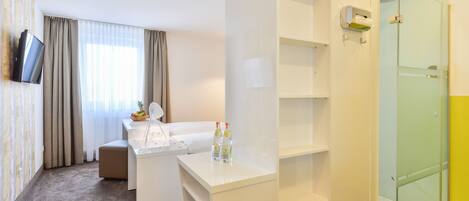A hotel room with a white desk, a TV mounted on the wall, a glass shower enclosure, and a window with curtains.