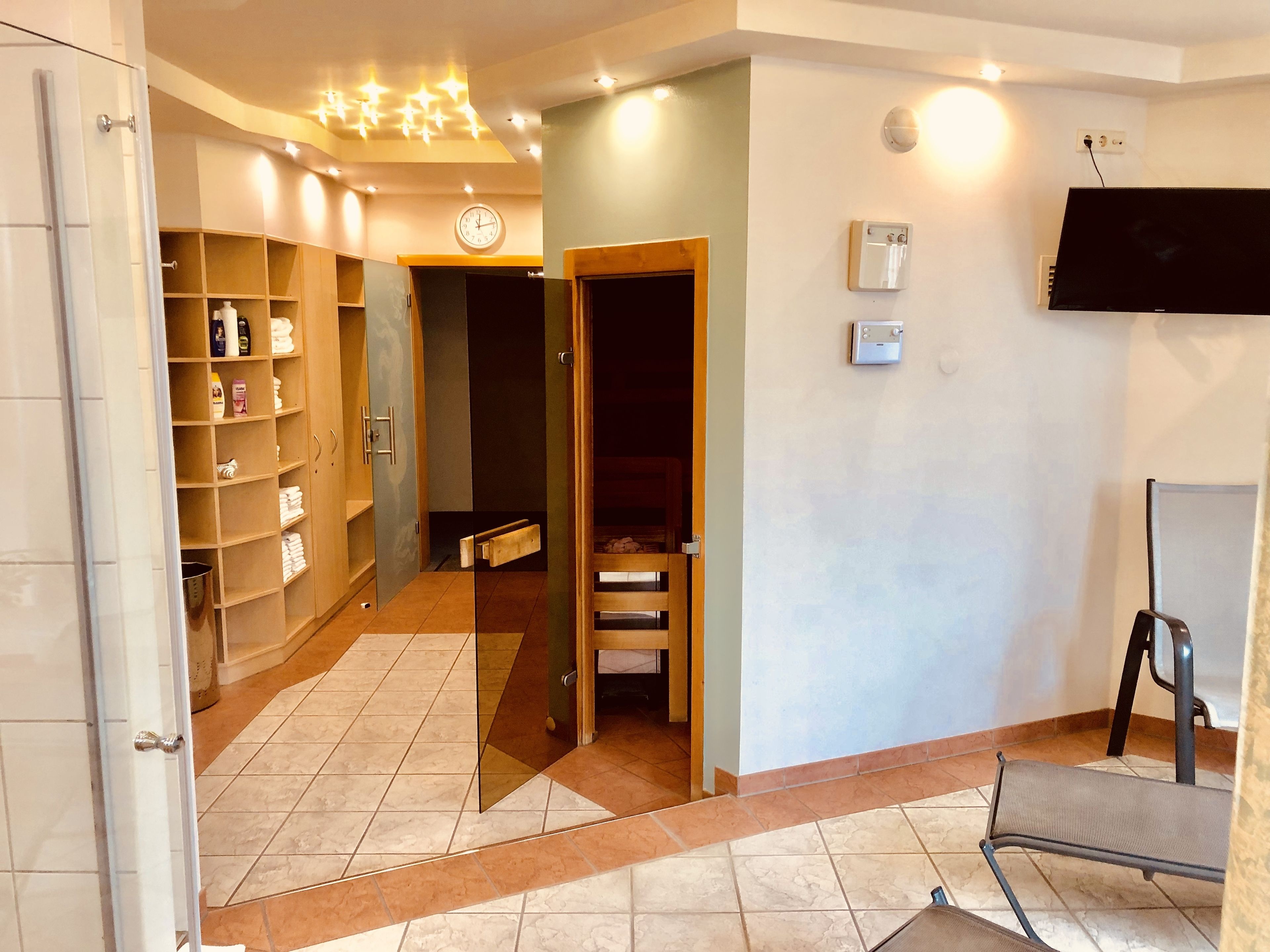 A hotel room with a tiled floor, a wooden wardrobe, a mirror, a clock, and a flat-screen TV mounted on the wall.