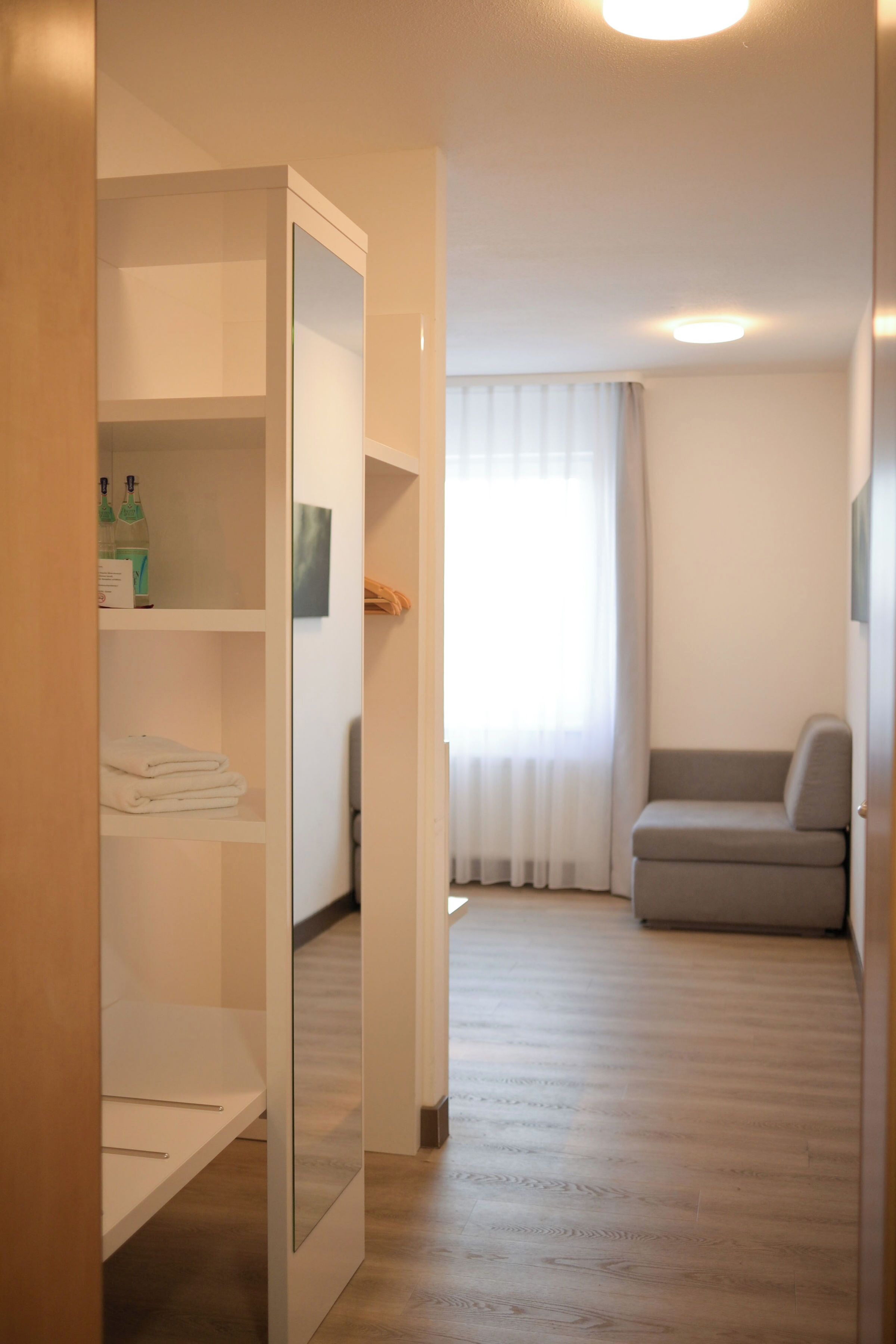 A room with a wardrobe, a grey sofa, a window with curtains, and a wooden floor.