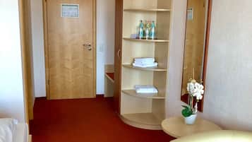 A hotel room with a red carpet, a wooden door, a wooden wardrobe with shelves, a white chair, a small table with a potted plant, and a mirror on the wall.