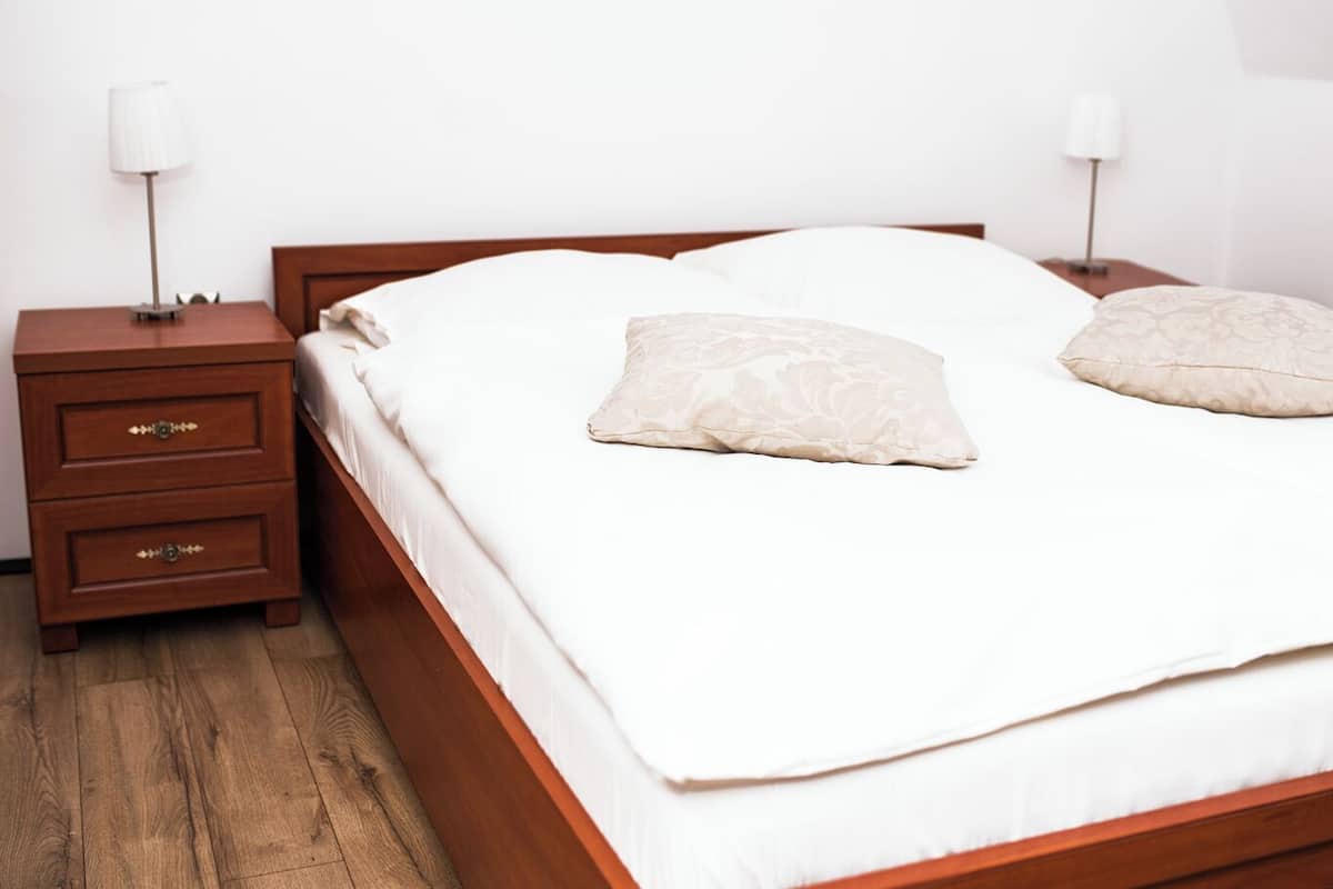 Een netjes opgemaakt bed met witte lakens en twee sierkussens, een nachtkastje met lades, en een bijzettafeltje met een lamp.