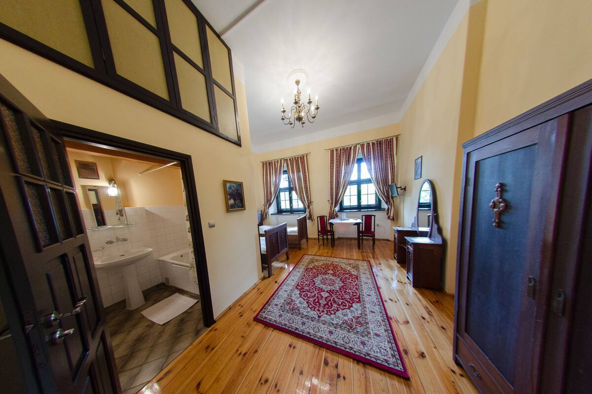 Een kamer met houten vloer, groot vloerkleed, een kroonluchter, bureau, stoel, kast en een badkamer met wastafel en bad.