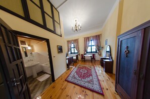 Una habitación con piso de madera, un tapiz grande, un candelabro, un escritorio, una silla, un armario y un baño con lavamanos y bañera.