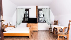 Un dormitorio con cama, televisor, cómoda, mesa con silla y dos ventanas con cortinas.