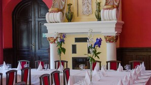 Una larga mesa de comedor cubierta con manteles, servilletas y copas blancas, flanqueada por sillas rojas, en una habitación con pared roja y puerta negra.