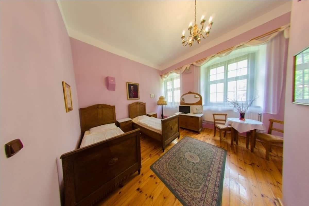 Een slaapkamer met twee houten bedden, een bureau, een stoel, een kroonluchter en een raam met gordijnen.