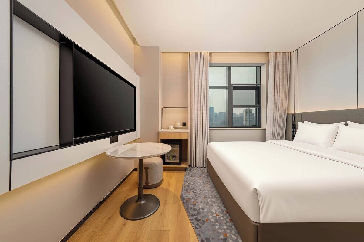 Ein Hotelzimmer mit Bett, Fernseher, einem kleinen Tisch mit Stuhl, einem Fenster mit Vorhängen und Blick auf die Stadt.