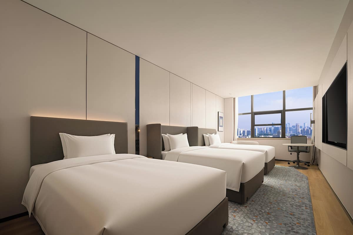 A hotel room with three beds, a desk, and a large window with a city view.