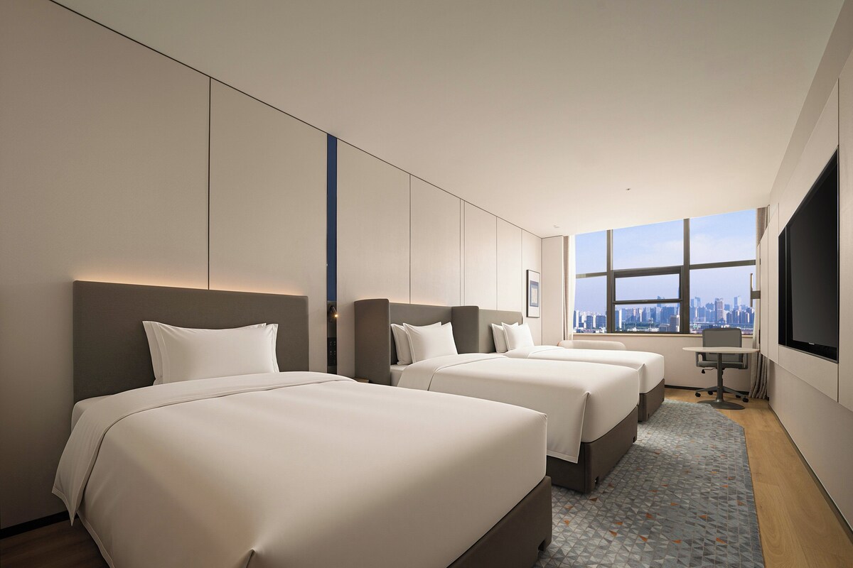 Ein Hotelzimmer mit drei Betten, einem Schreibtisch und einem großen Fenster mit Blick auf die Stadt.