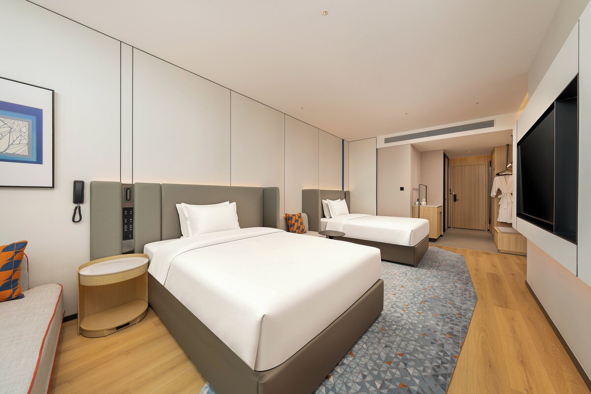 A modern hotel room with a large bed, a bedside table, a chair, a desk, a mirror, and a TV.