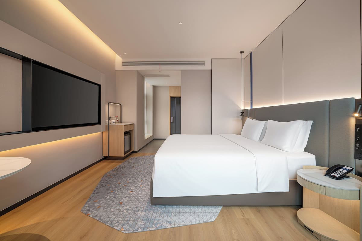 A modern hotel room with a large bed, bedside table, built-in TV, and a small desk with a phone.
