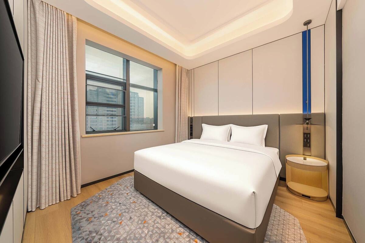 Ein modernes Hotelzimmer mit einem großen Bett, einem Fenster mit Vorhängen, einem Nachttisch und einer Lampe.