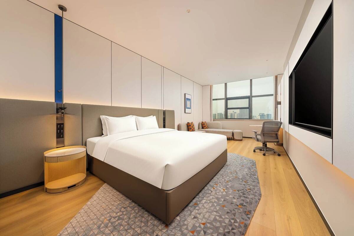 Ein modernes Hotelzimmer mit einem großen Bett, einem Schreibtisch, einem Stuhl, einem Fernseher und einem Fenster mit Blick auf die Stadt.