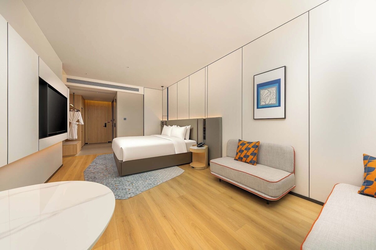 A modern hotel room with a bed, sofa, TV, and artwork.