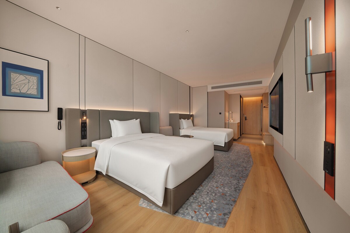 A modern hotel room with a large bed, a smaller bed, a sofa, a bedside table, and a TV.