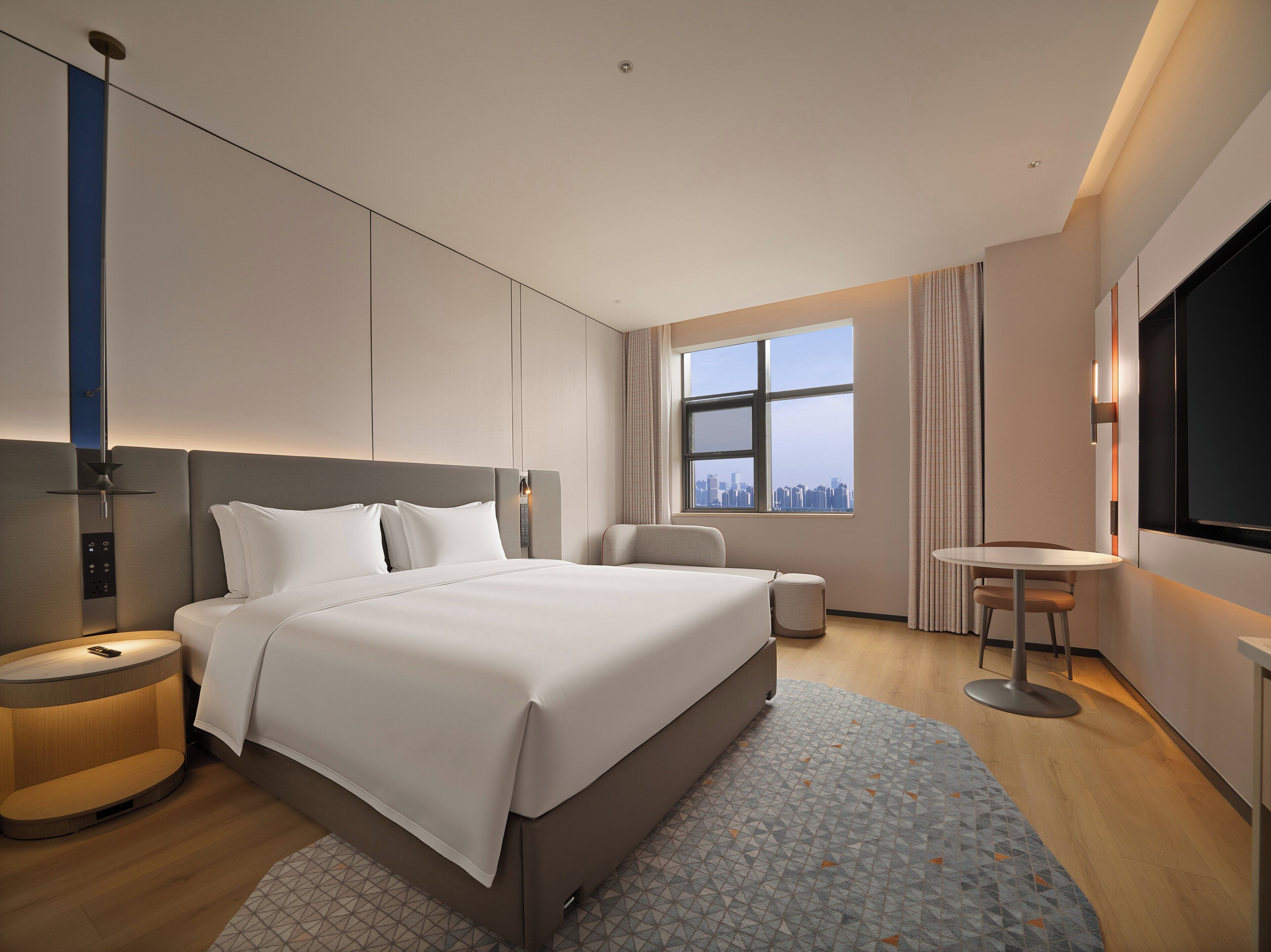 A modern hotel room with a large bed, bedside tables, a desk, and a city view.
