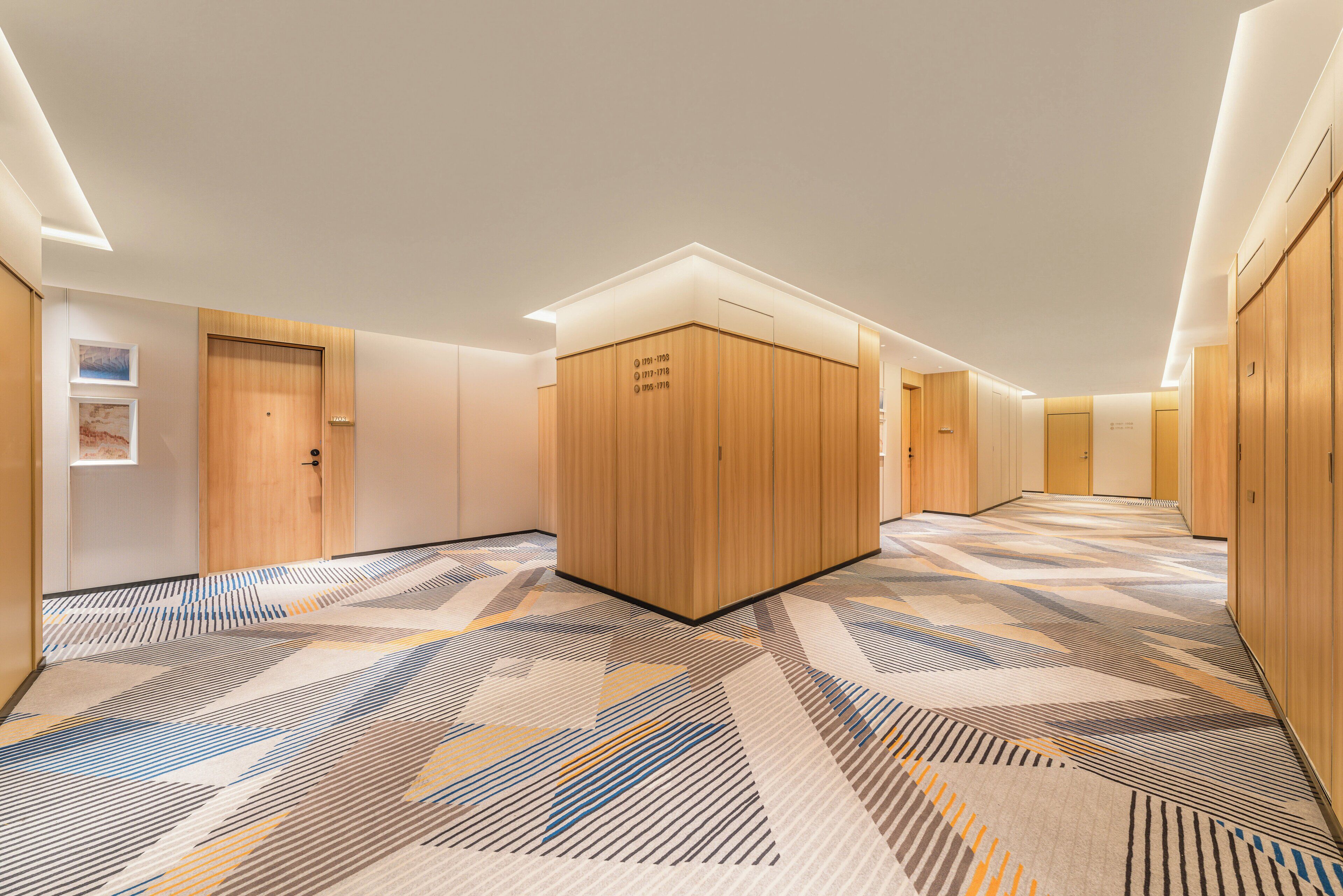 Pasillo de hotel con puertas de madera, alfombra estampada e iluminación empotrada.