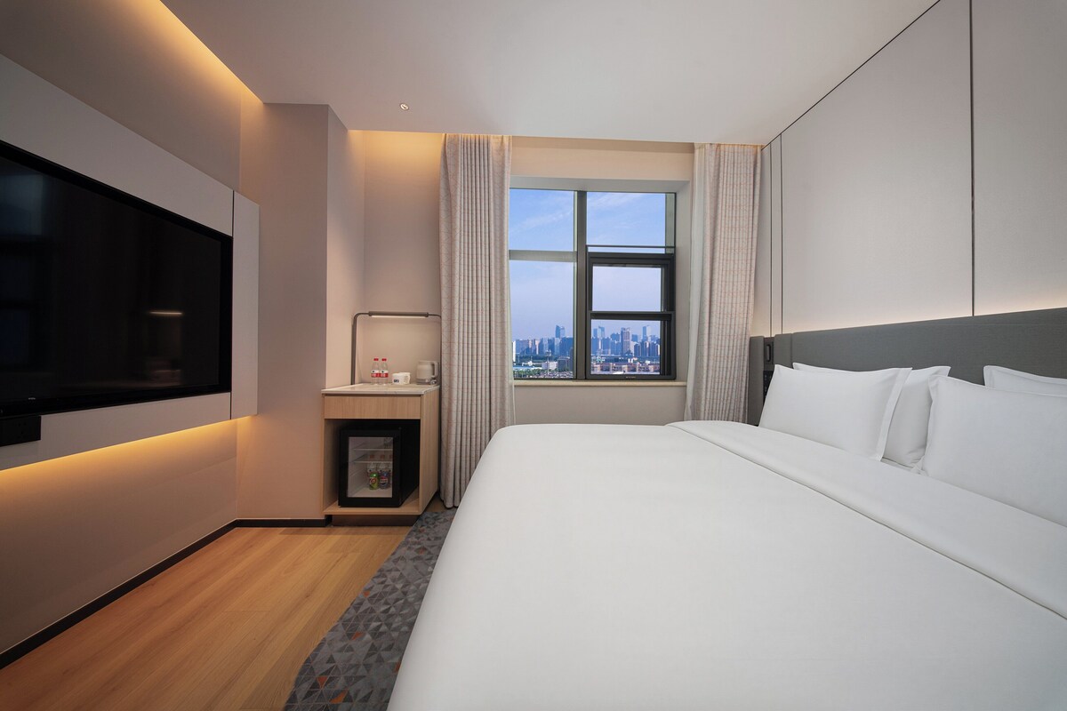 Ein Hotelzimmer mit einem großen Bett, einem Fernseher, einem Schreibtisch mit Lampe, einem Fenster mit Stadtblick und einer kleinen Ablagefläche.