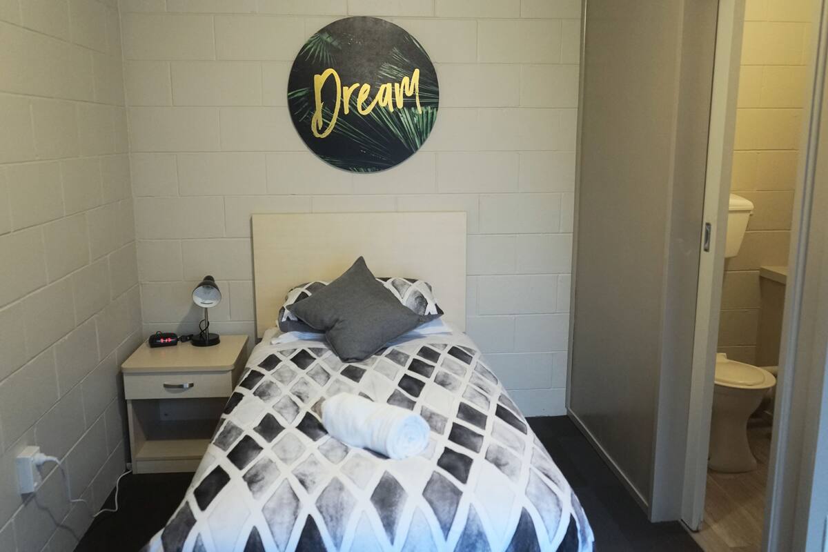 A small, neatly arranged bedroom with a bed, a bedside table with a lamp, and a wall-mounted sign that reads "Dream".