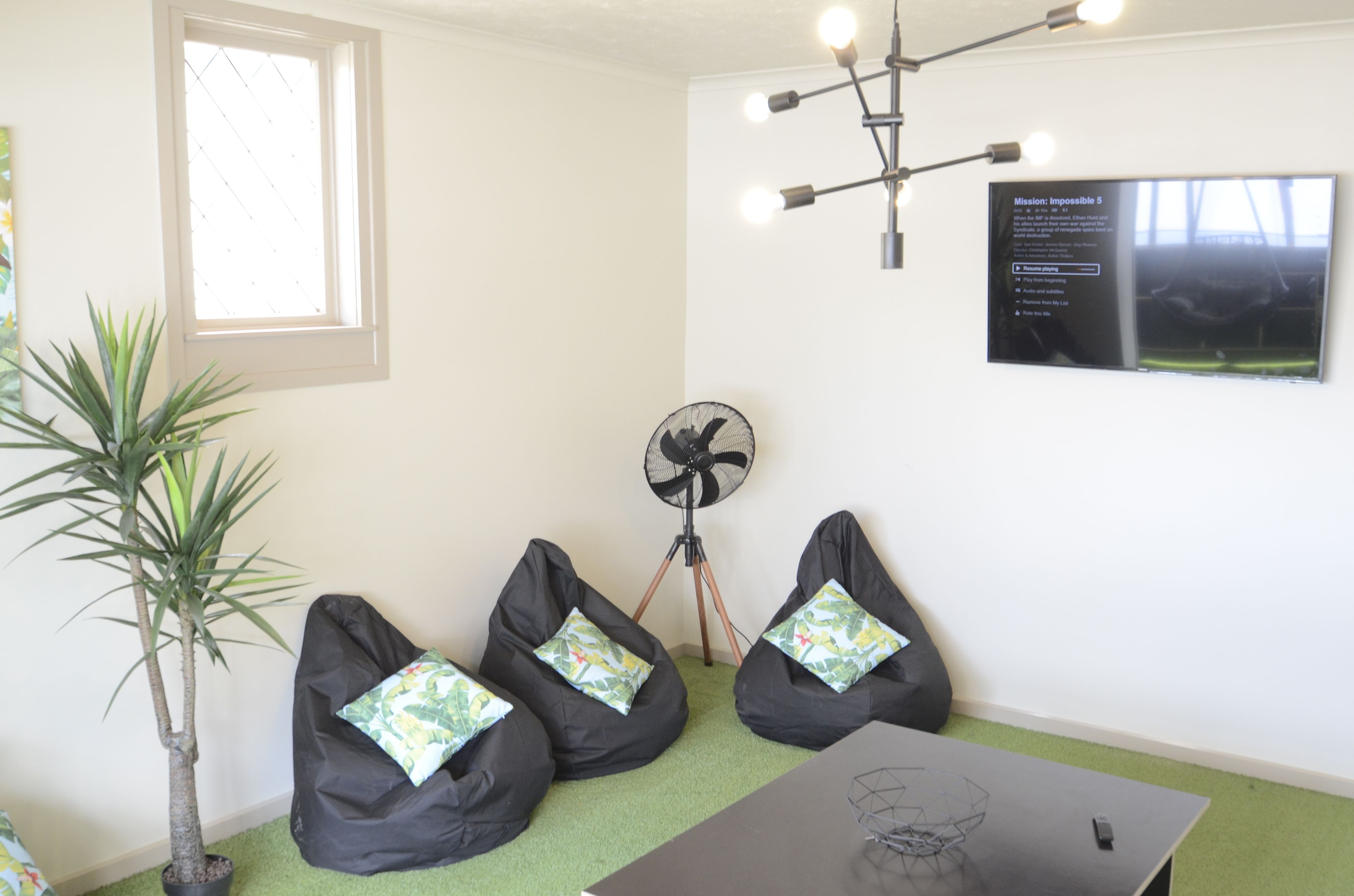 A room with three black bean bags, a standing fan, a potted plant, and a mounted TV displaying a message.
