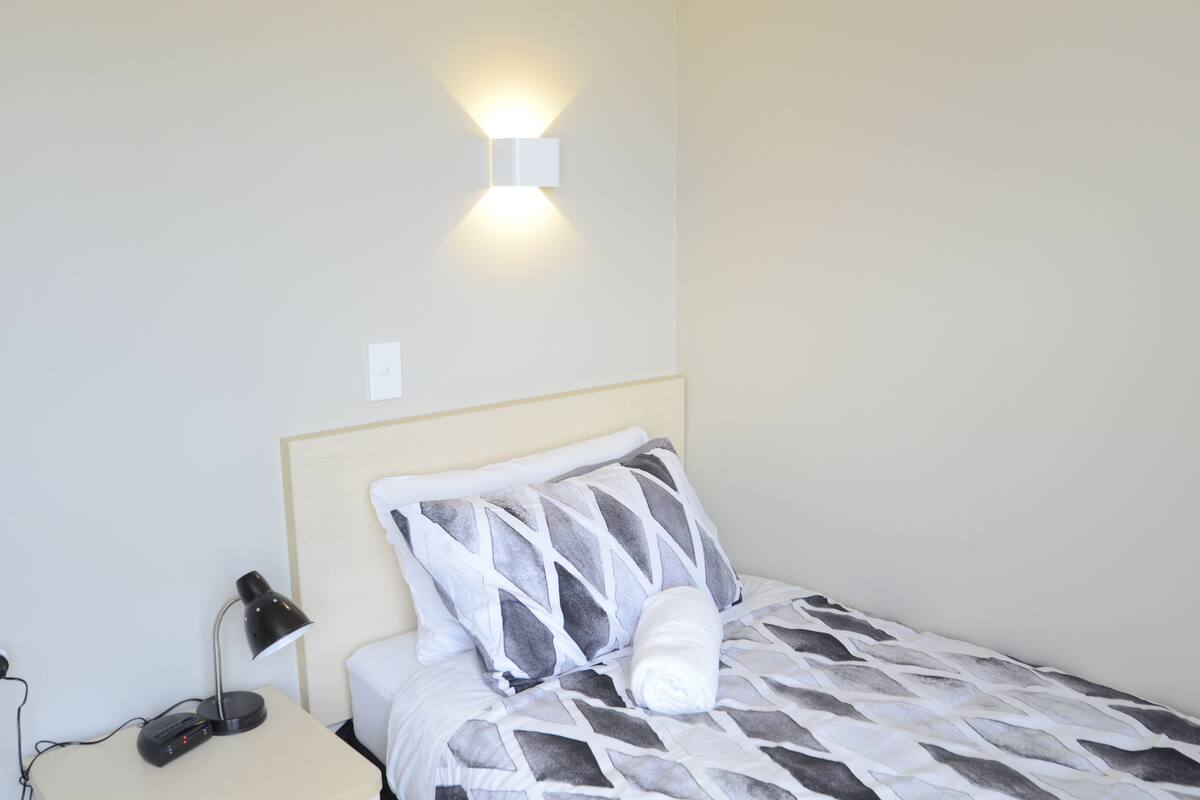 A neatly made bed with a patterned bedspread, a white pillow, and a rolled-up towel. There is a black desk lamp on a bedside table, and a wall-mounted light fixture above the bed.