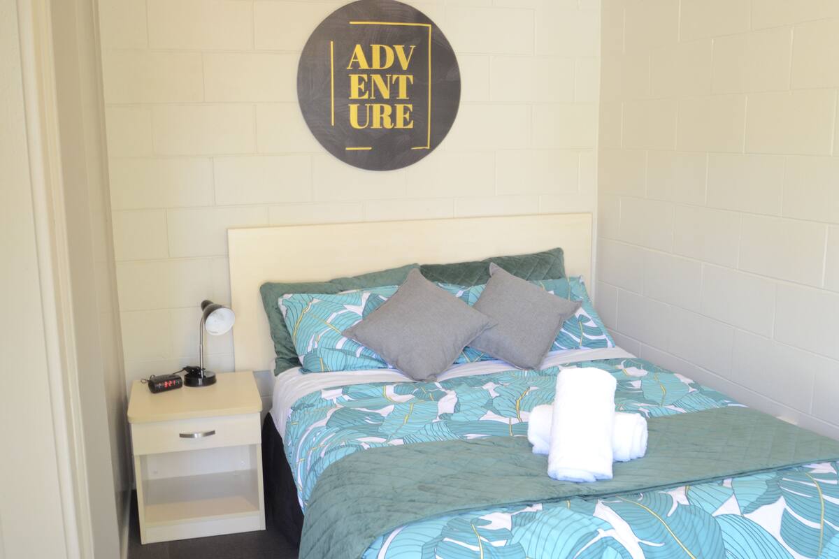 A neatly made bed with a patterned bedspread, two pillows, a bedside lamp, and a wall-mounted sign that reads "ADVENTURE".