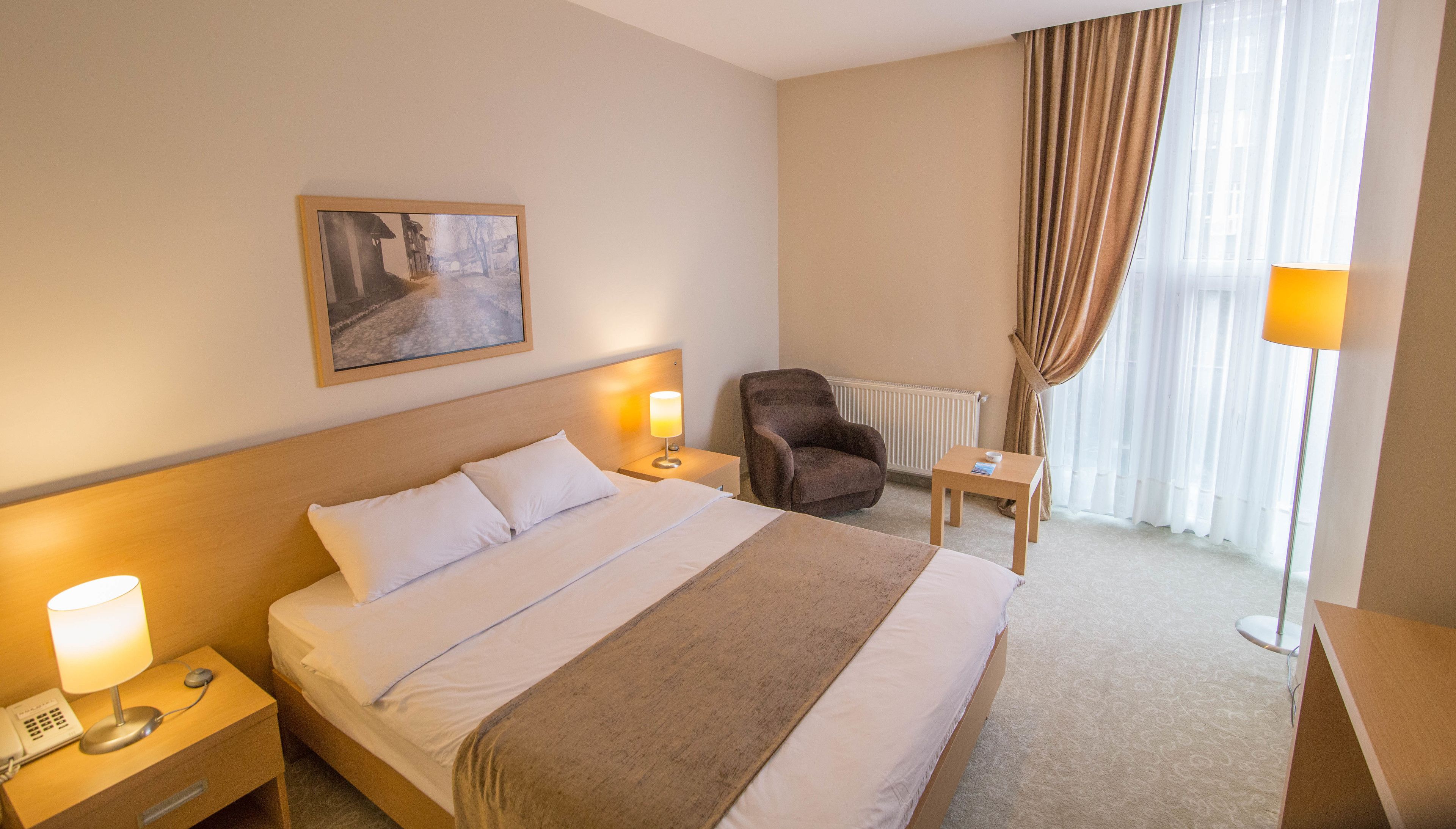 Une chambre d’hôtel avec un lit, des lampes de chevet, une chaise, une petite table, un radiateur et une fenêtre avec des rideaux.