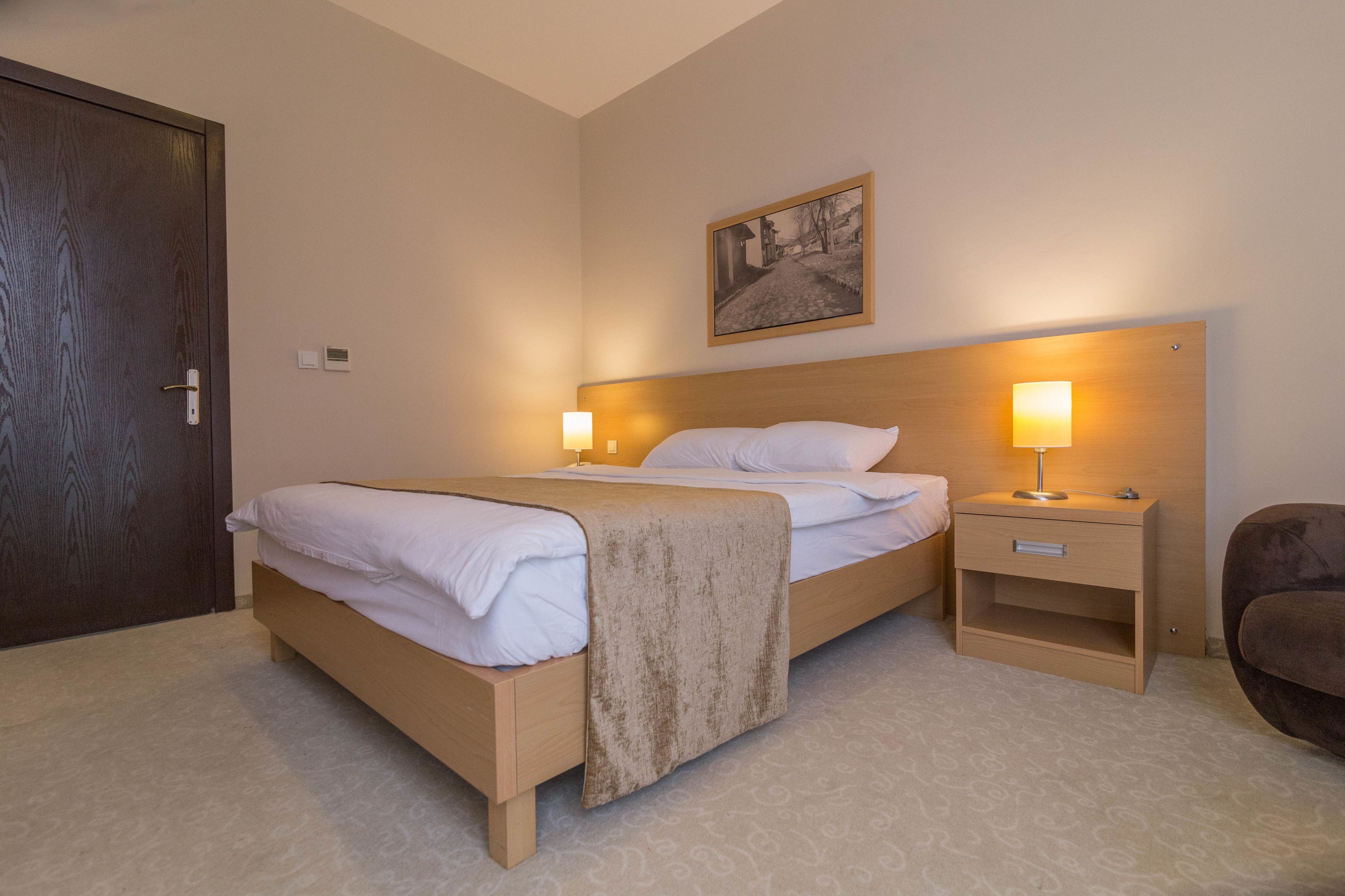Quarto de hotel com cama de madeira, mesas de cabeceira com abajures, uma cadeira e um quadro na parede.