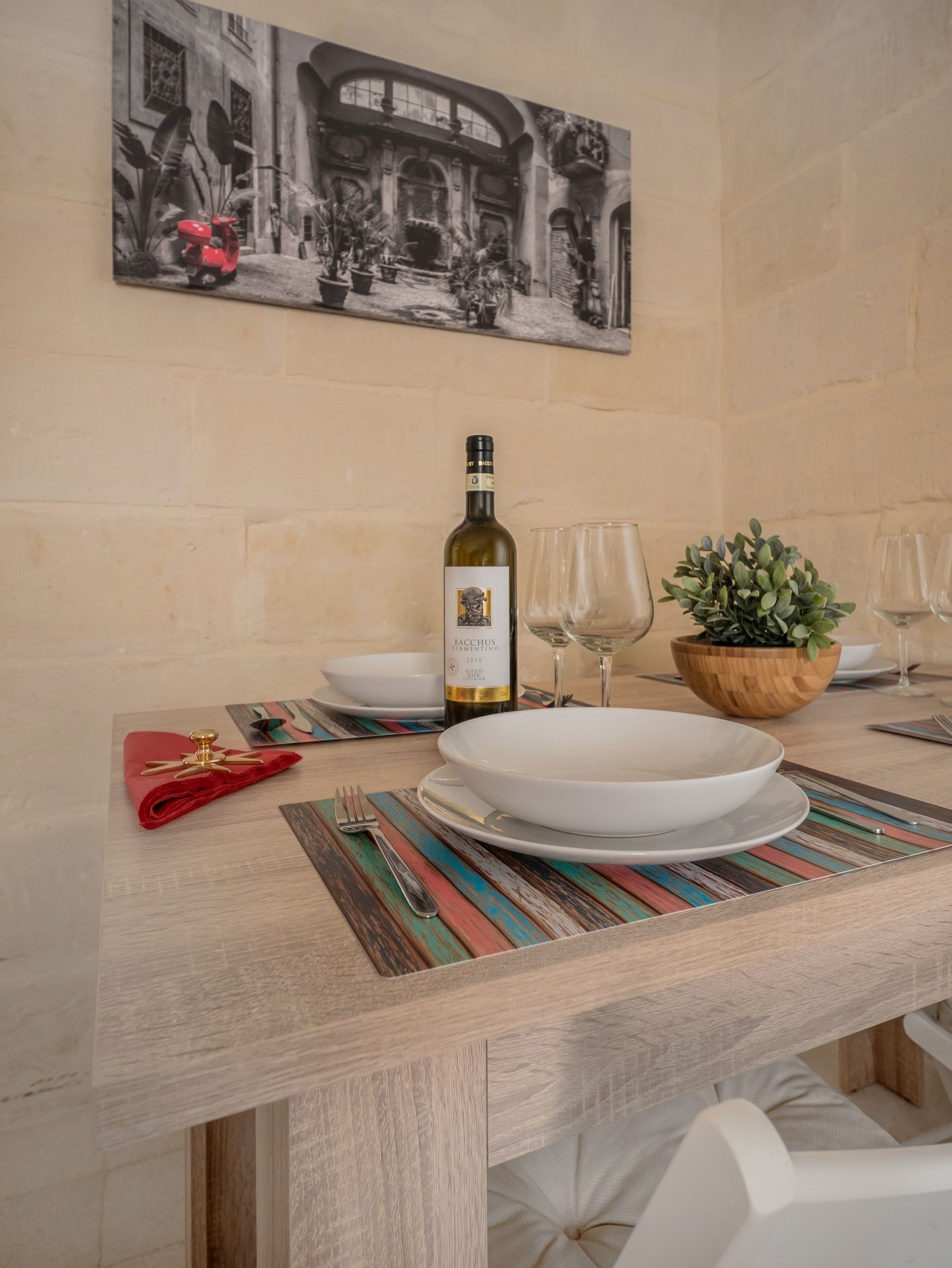 Una mesa de comedor dispuesta con una botella de vino, copas, platos y un mantel individual rojo.