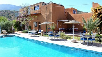 A pool area with lounge chairs and umbrellas, a building with a flat roof, and a mountainous backdrop.