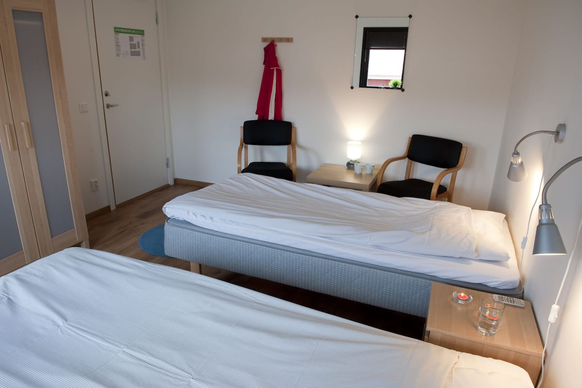 A hotel room with two single beds, a small table with a lamp, two chairs, a mirror, and a door with a sign.