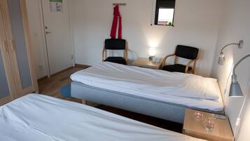 Habitación de hotel con dos camas individuales, una mesita con lámpara, dos sillas, un espejo y una puerta con un cartel.