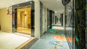 A hotel corridor with a patterned carpet, a glass elevator, and decorative wall panels. - Lvgu Hotel Yiwu (Jinhua)