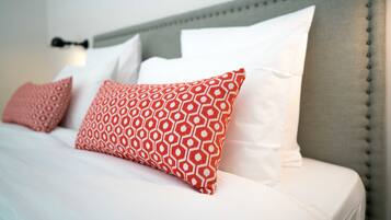 Ein ordentlich bezogenes Bett mit weißen und roten Kissen, ein grauer möbelbezogener Kopfteil und eine wandmontierte Leuchte.