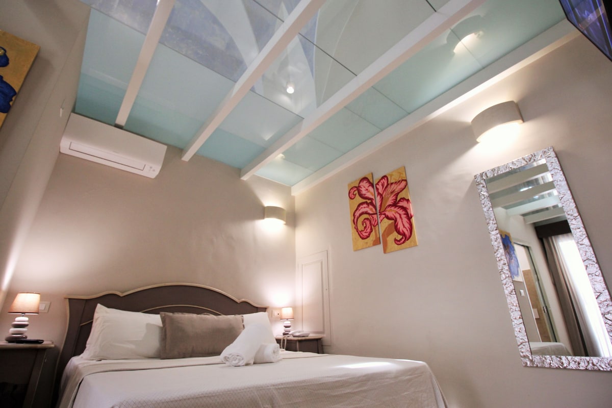 Habitación de hotel con cama, mesita de noche, espejo en la pared y techo con tragaluces.