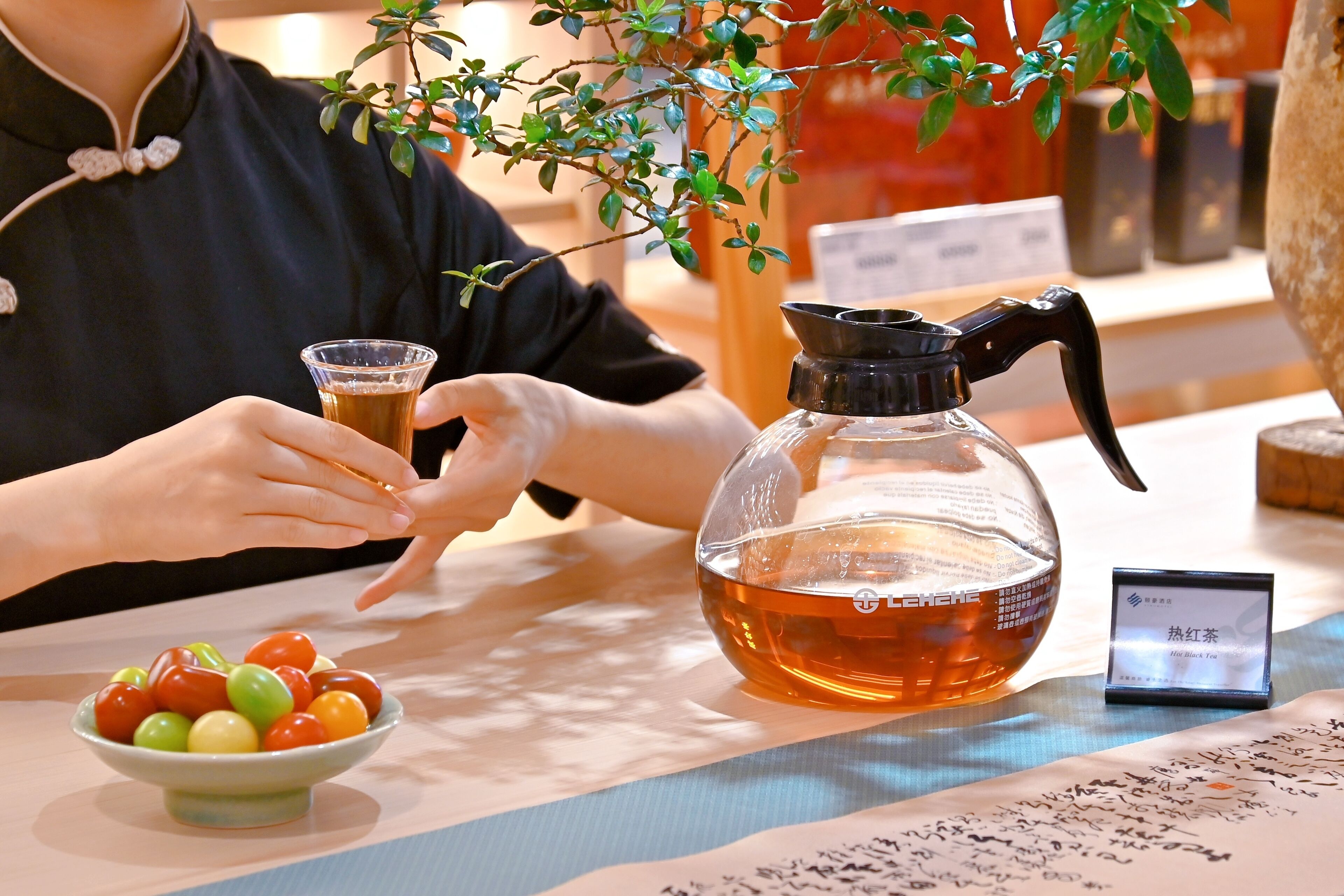A person holding a glass of tea, a teapot, and a bowl of cherry tomatoes on a table with a plant in the background.