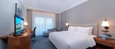 A hotel room with a large bed, a desk with a computer, a chair, a small table with a lamp, and a window with curtains.