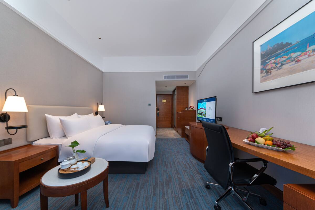 Ein Hotelzimmer mit einem Bett, Nachttischen, einem Schreibtisch mit Computer, einem Sessel, einem kleinen Tisch mit Früchten und einem Bild an der Wand.