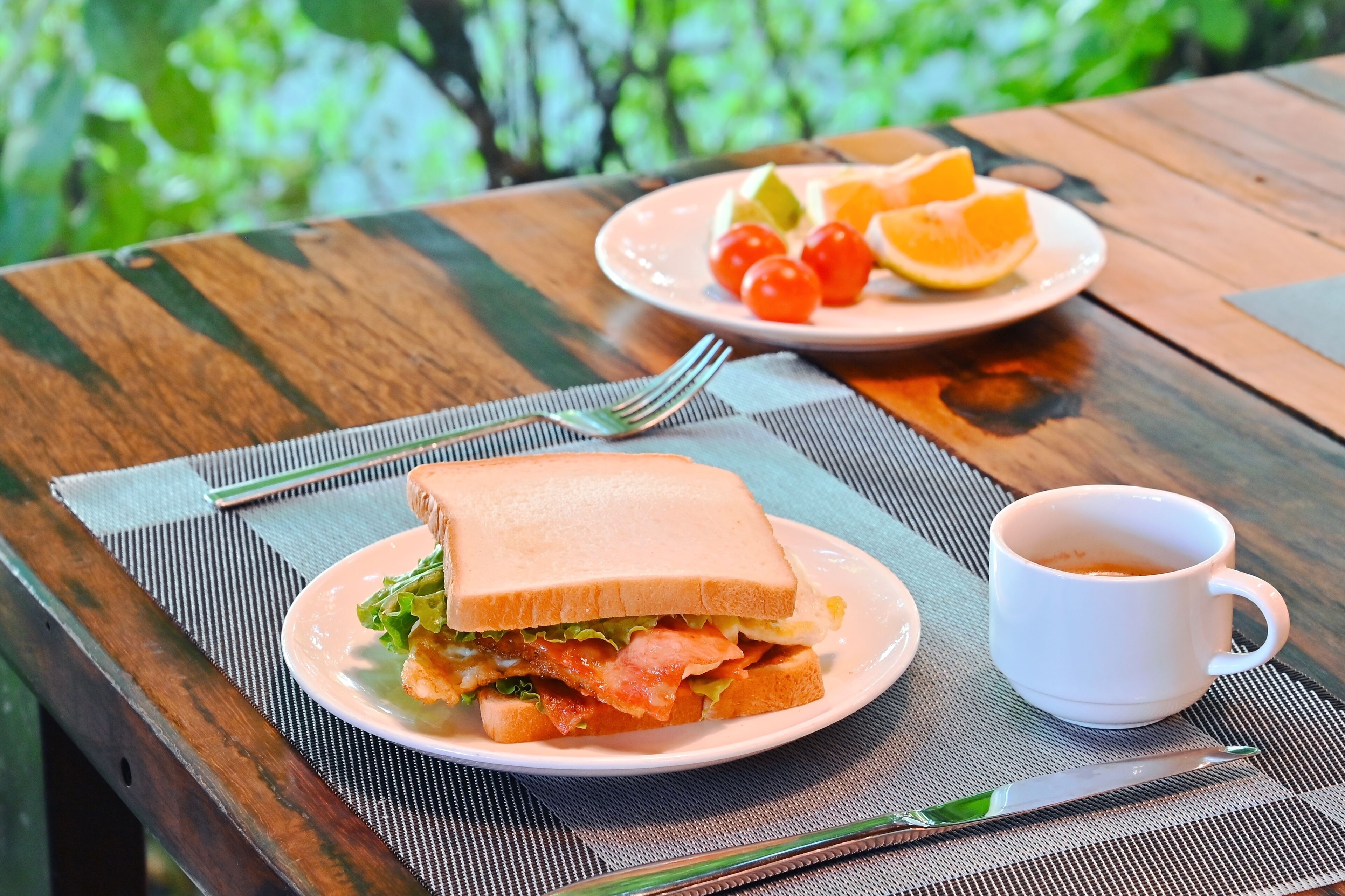 A wooden table with a plate of sandwich, a cup of coffee, and a plate of fruit.