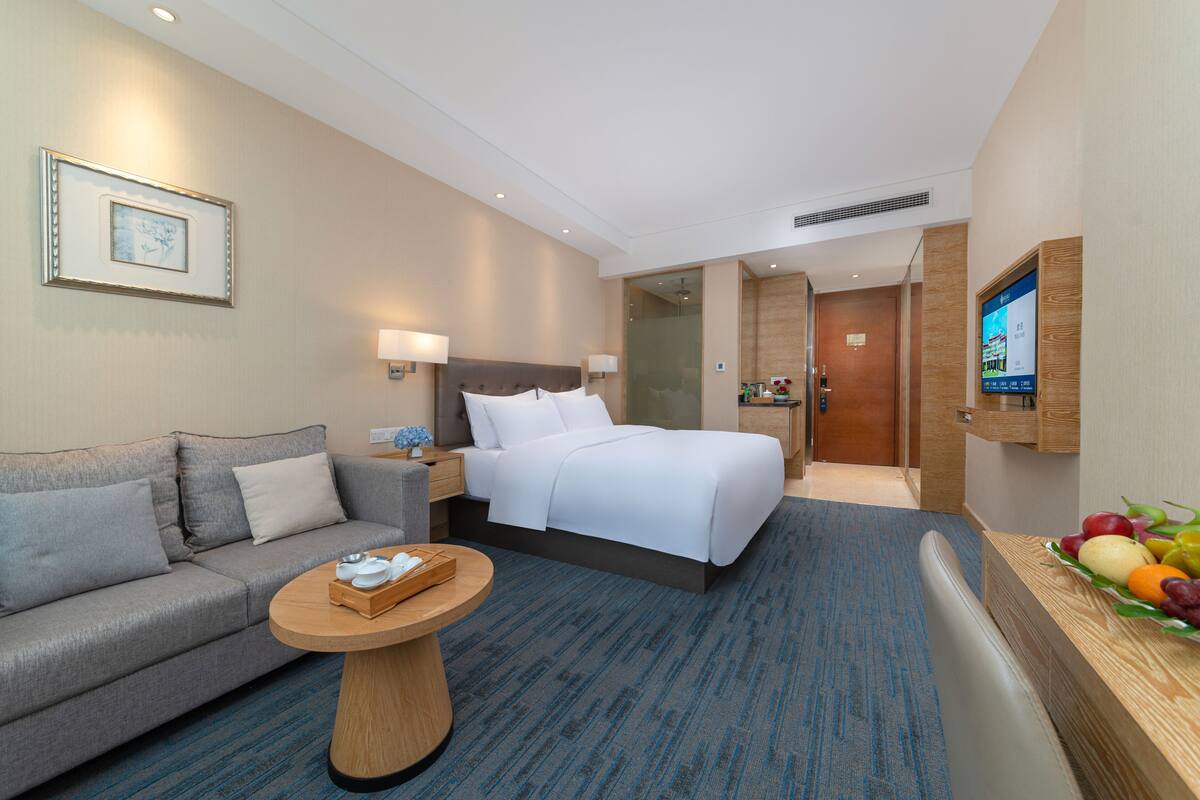 Ein Hotelzimmer mit einem Bett, einem Sofa, einem kleinen runden Tisch mit Tablett, einem an der Wand befestigten Fernseher und einer Obstschale auf dem Nachttisch.