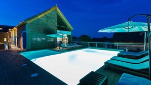 Un espace piscine extérieur moderne, doté de chaises longues et de parasols, entouré d’une terrasse en bois et d’un bâtiment au toit en pente.