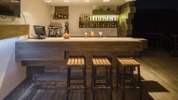 Zona de bar con mostrador de madera, taburetes, una máquina de café y varias botellas en estanterías.