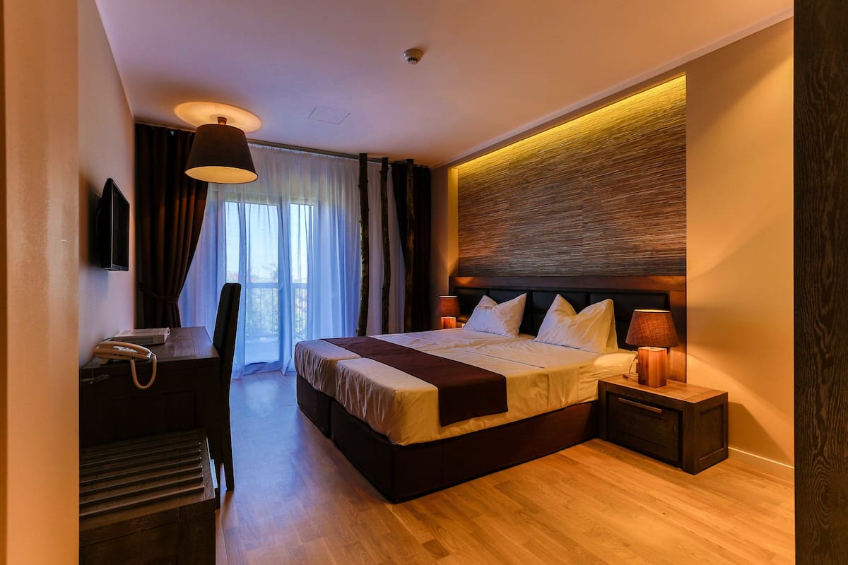 Ein modernes Hotelzimmer mit einem großen Bett, Nachttischlampen, einem Schreibtisch mit Telefon und Blick ins Freie durch das Fenster.
