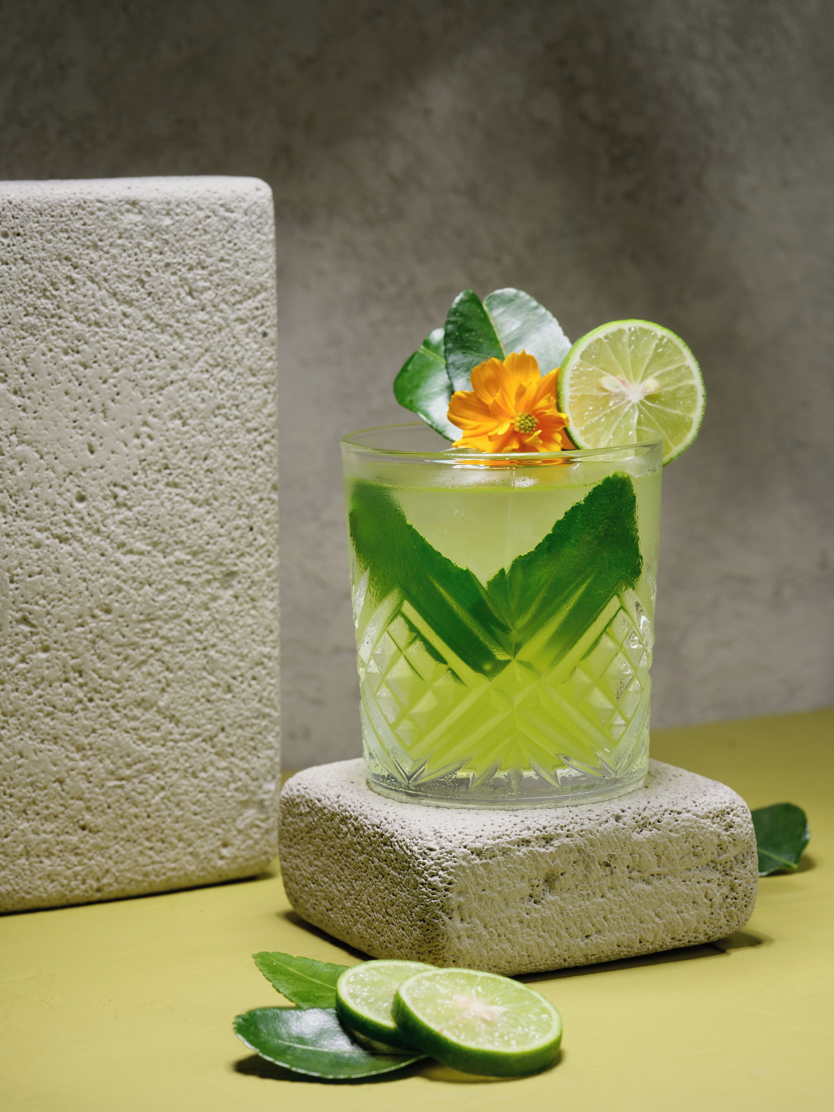 A glass of green beverage with lime slices and a flower, placed on a textured stone.