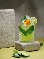 A glass of green beverage with lime slices and a flower, placed on a textured stone.