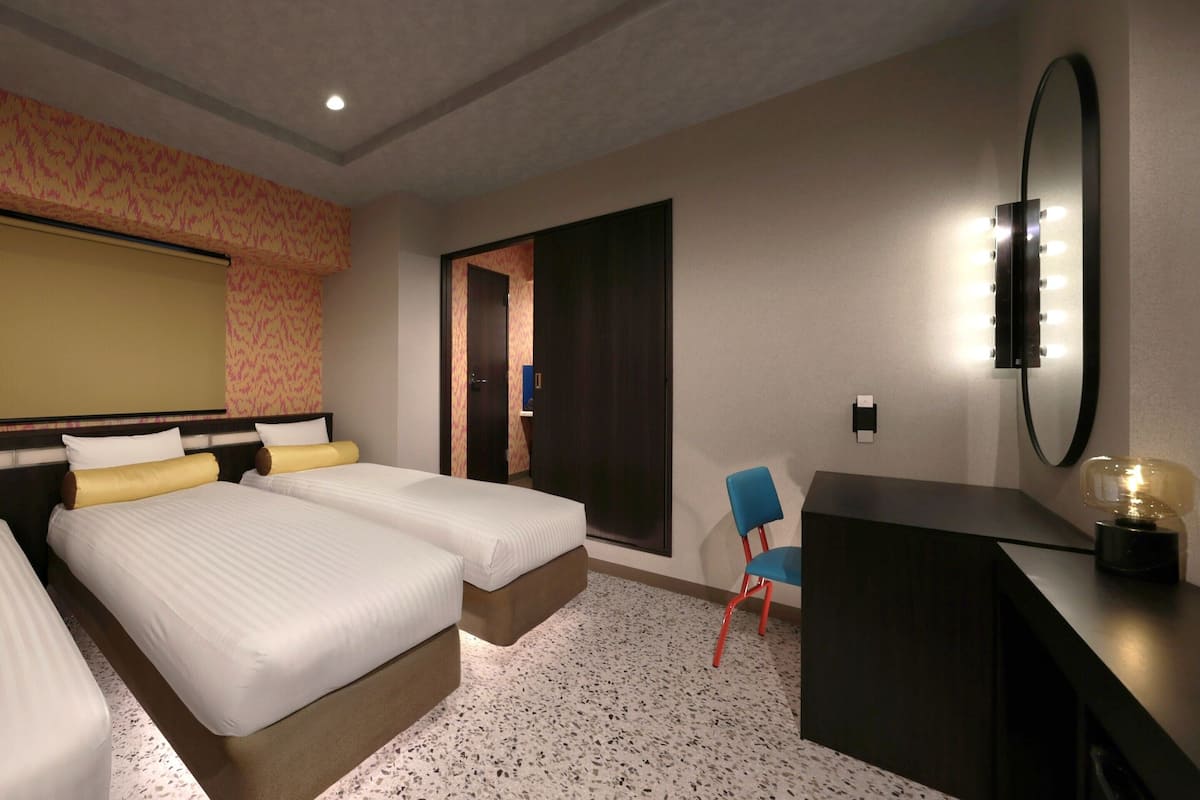 A hotel room with two beds, a desk, a chair, a mirror with lights, and a door leading to another room.