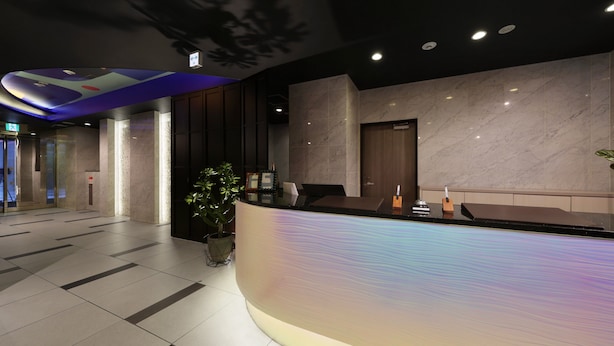 A modern reception area with a curved counter, marble walls, and a potted plant. - Nipponbashi LUXE Hotel (Osaka)