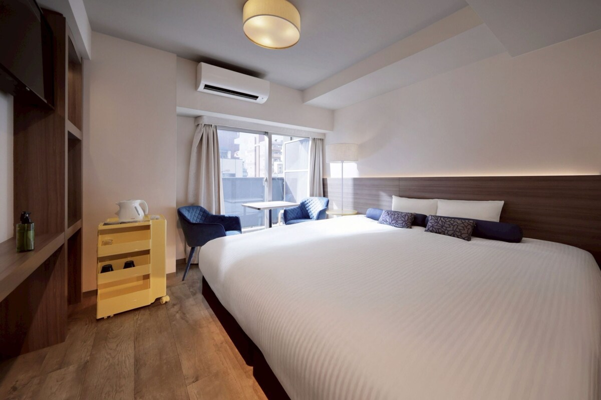 Habitación de hotel con una cama grande, una mesita, una silla y una lámpara de noche.